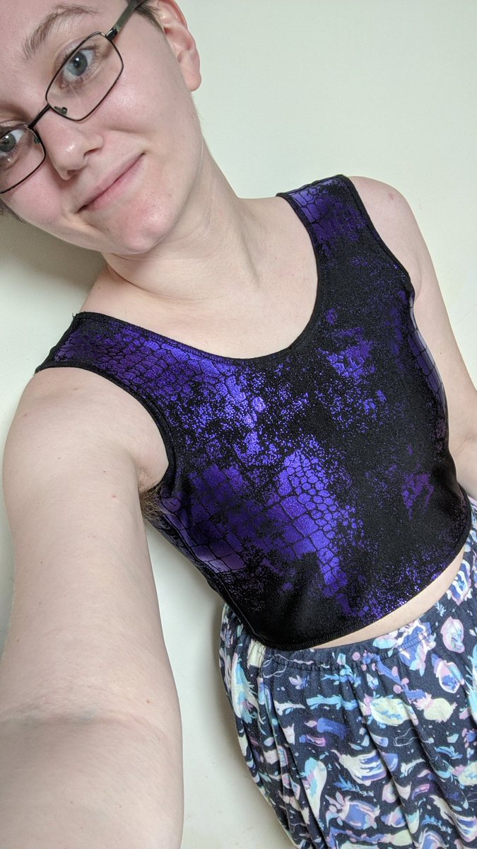 A white demiboy with thin black glasses and buzzed hair is taking a selfie of themself. They are wearing a crop-top length purple snake-print chest binder and a purple skirt with crystals, mushrooms, jars, succulents, and other witchy things printed onto it.