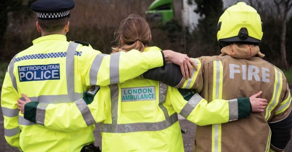 Three emergency service personnel stand next to each other, arm in arm. From left to right there appears a London Met policeman, a London ambulance worker and a London fire brigade member of staff. 