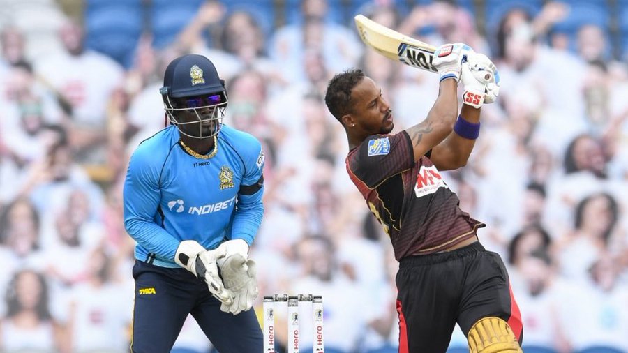 St Lucia Zouks wicketkeeper Andre Fletcher watches on as Trinbago Knight Riders batter Lendl Simmons hits a powerful shot. The Zouks shirt is sea blue with a yellow collar and the trousers are navy blue. Fletcher is wearing a dark blue helmet and white wicketkeeper’s gloves. The Knight Riders shirt is dark red with black sleeves and side panels and gold trim. The trousers are black with thick red piping. Simmons is bare-headed and wearing white batting gloves and gold batting pads.