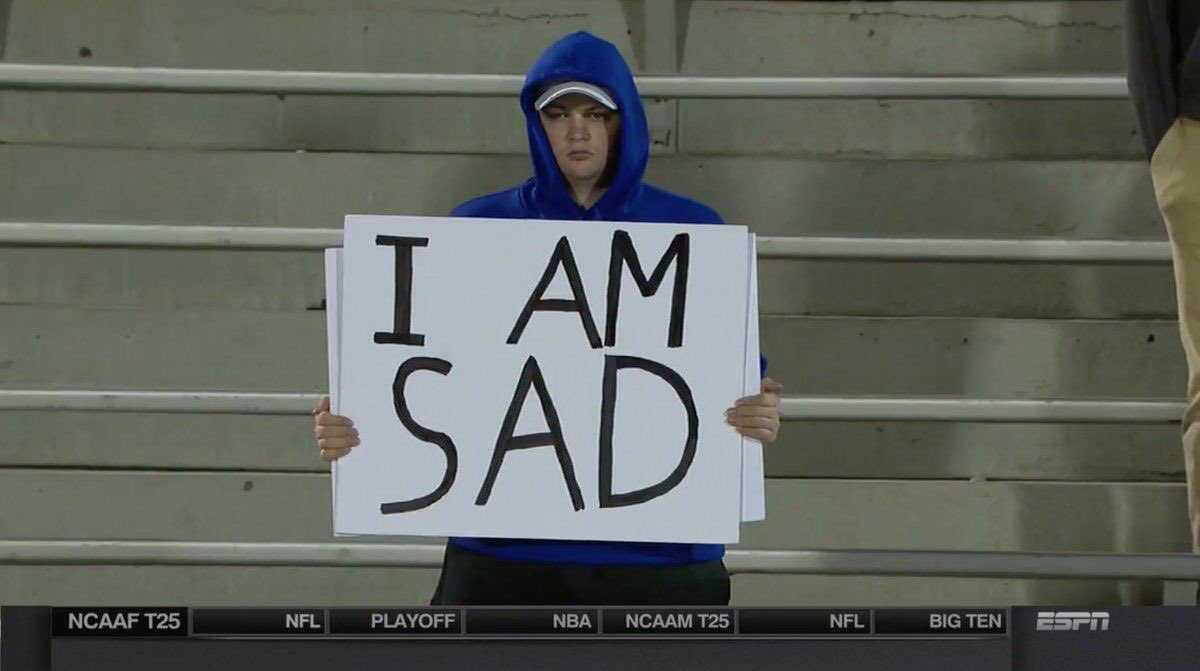 A football fan standing alone in the stadium holding a sign that reads "I AM SAD".