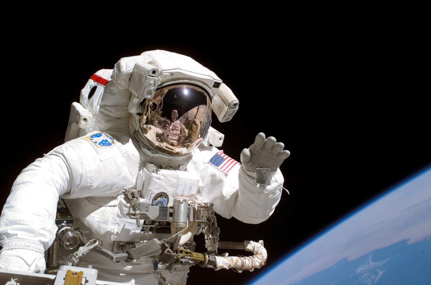 Astronaut in spacesuit on a spacewalk. Pitch black space can be seen behind him, with a sliver of Earth in the bottom right corner.