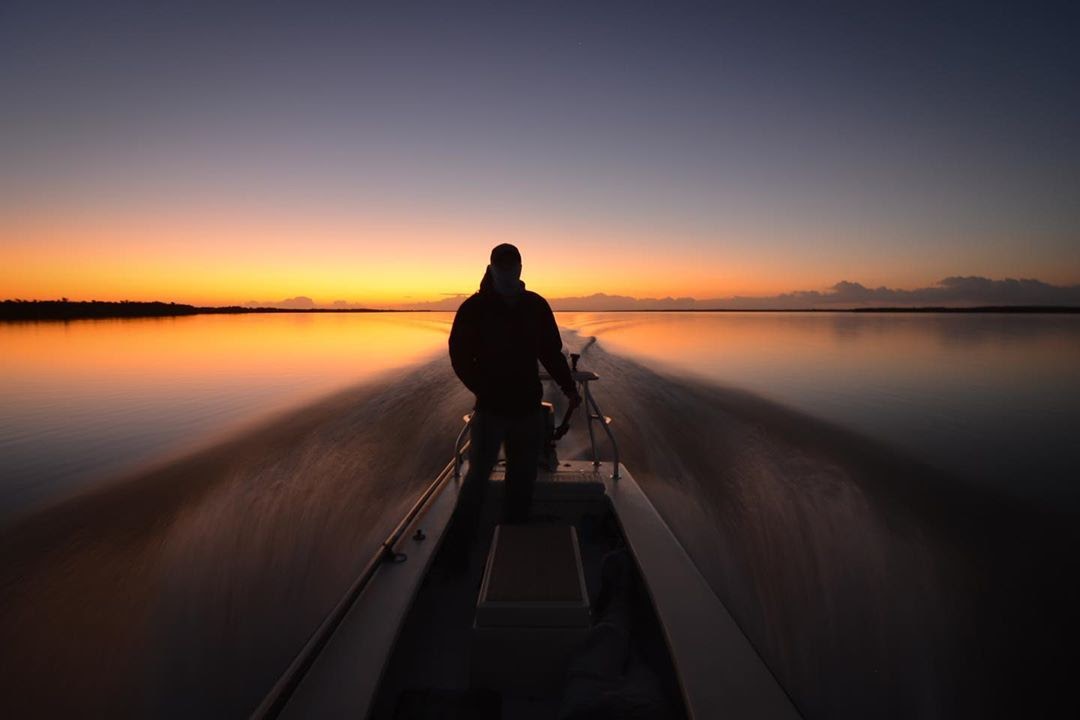 Just before sunrise on the water. At the horizon the night sky begins to turn from grey to fiery hues of purple, pink, and orange. A person stands holding the tiller pole on an outboard motorboat speeding away from the horizon.