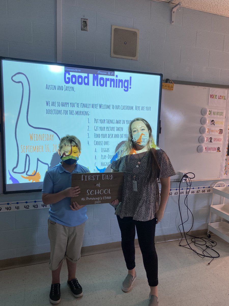 male student and female teacher, wearing masks, pose for first day of school photo in classroom.
