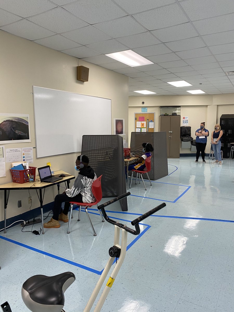 two students, a boy and a girl, sit at computers doing school work. they are socially distanced, wearing masks. two teachers stand in the distance.