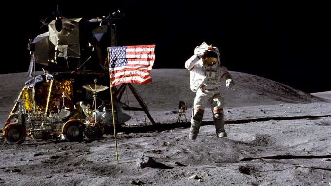 Astronaut in spacesuit salutes American flag on the Moon; lunar module and lunar roving vehicle are in the background 