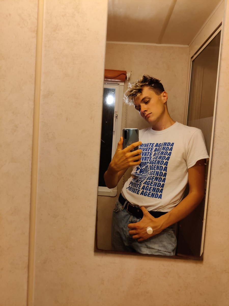 Selfie in caravan bathroom mirror, wearing blue jeans, navy belt and whit t shirt with blue text that reads 'Private Agenda'