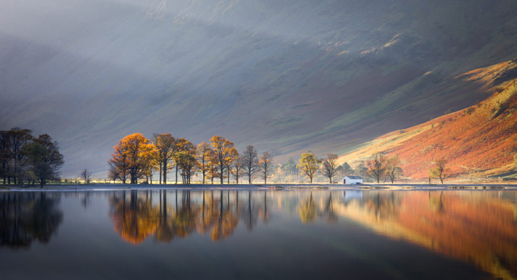 A house by a lake at the bottom of a mountain covered in autumnal colours