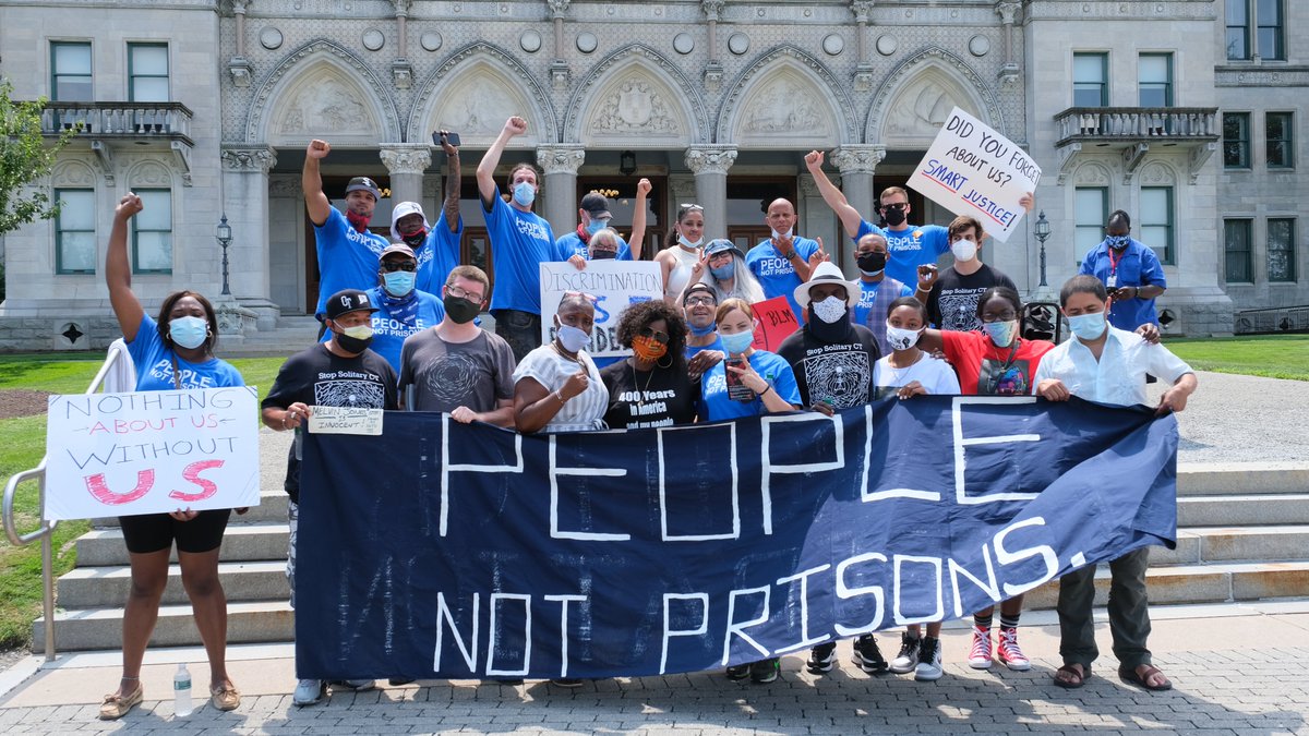 A group of people in masks hold a navy blue "people not prisons" banner. They are standing, outside of the Connecticut State Capitol Building, some with signs and some with fists up. Most are wearing bright blue "people not prisons" ACLU-CT Smart Justice shirts.