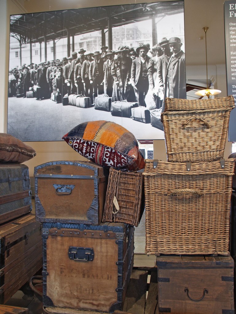 Historic photo of immigrants waiting in line with luggage in background, with replica luggage featuring baskets, pillows, and chests in forefront. 
