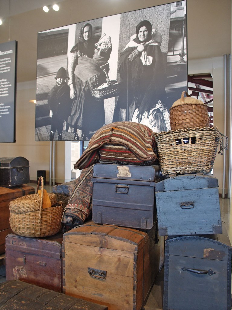Replica luggage featuring chests, baskets, and pillows in forefront with an historic image of immigrant family in background. 