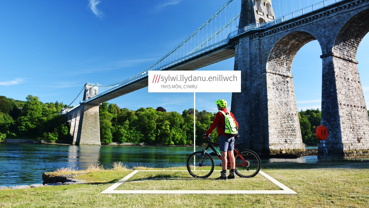 Cyclist next to river, bridge in the background. what3words address is in welsh and reads ///sylwi.llydanu.enillwch