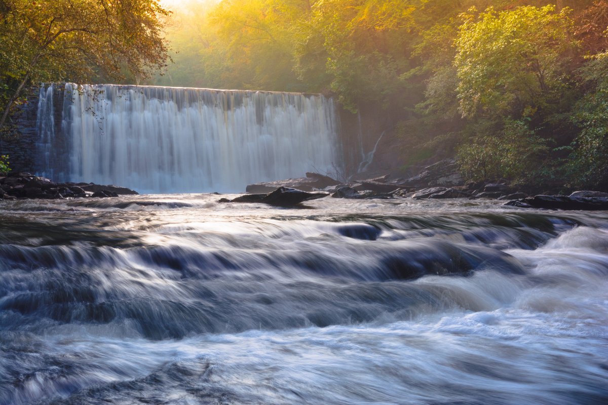 A water fall flows into a river bordered by trees showing fall colors.