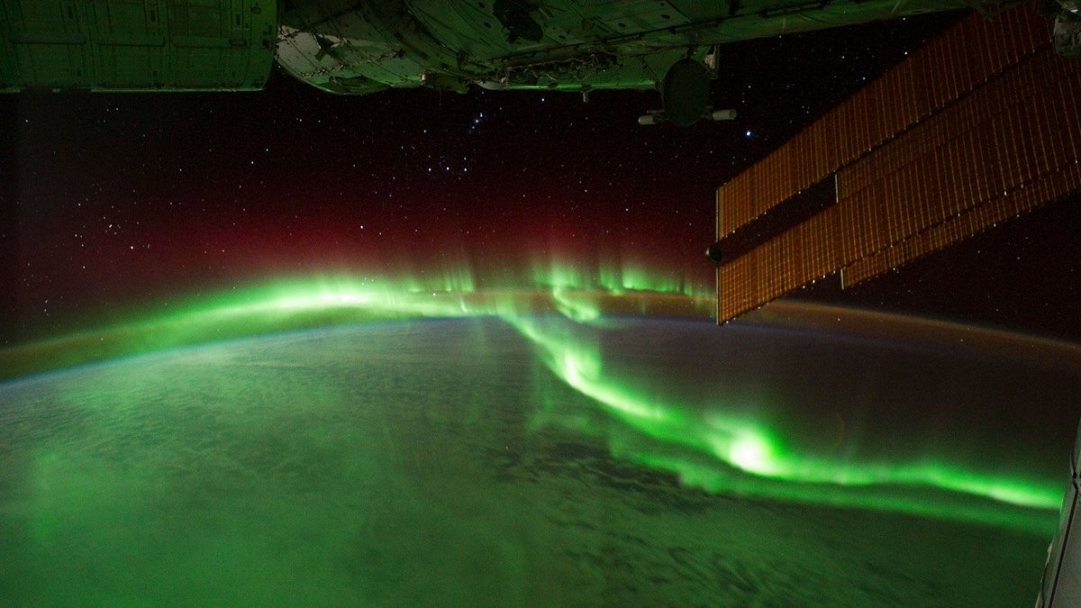 View of Earth from a satellite orbiting in space. Green auroras are lighting up Earth' surface in the lower half of the frame. Dark space makes up the upper half of the frame; stars can be seen scattered throughout. 