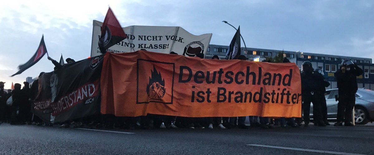 Fronttransparent "Deutschland ist Brandstifter" dahinter der Demozug. Schwarze und rote Fahnen.