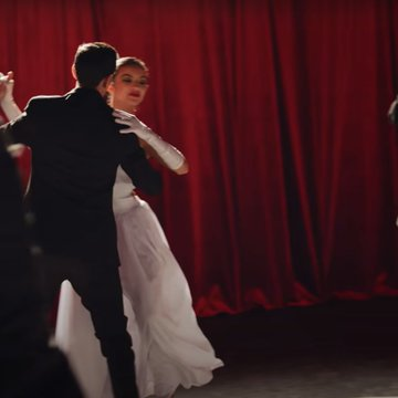 The dancing couples with their outfits and the air of a formal dance remind me of debutante balls, which are about making a debut in the world and showing off young adults in high society - it's not really a loving/fun dance. He's just waiting in a world of pretenses.