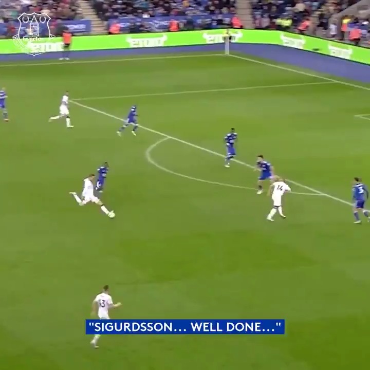 A wide shot of a soccer match in progress on a green field, featuring the text overlay 'SIGURDSSON... WELL DONE...' at the bottom.