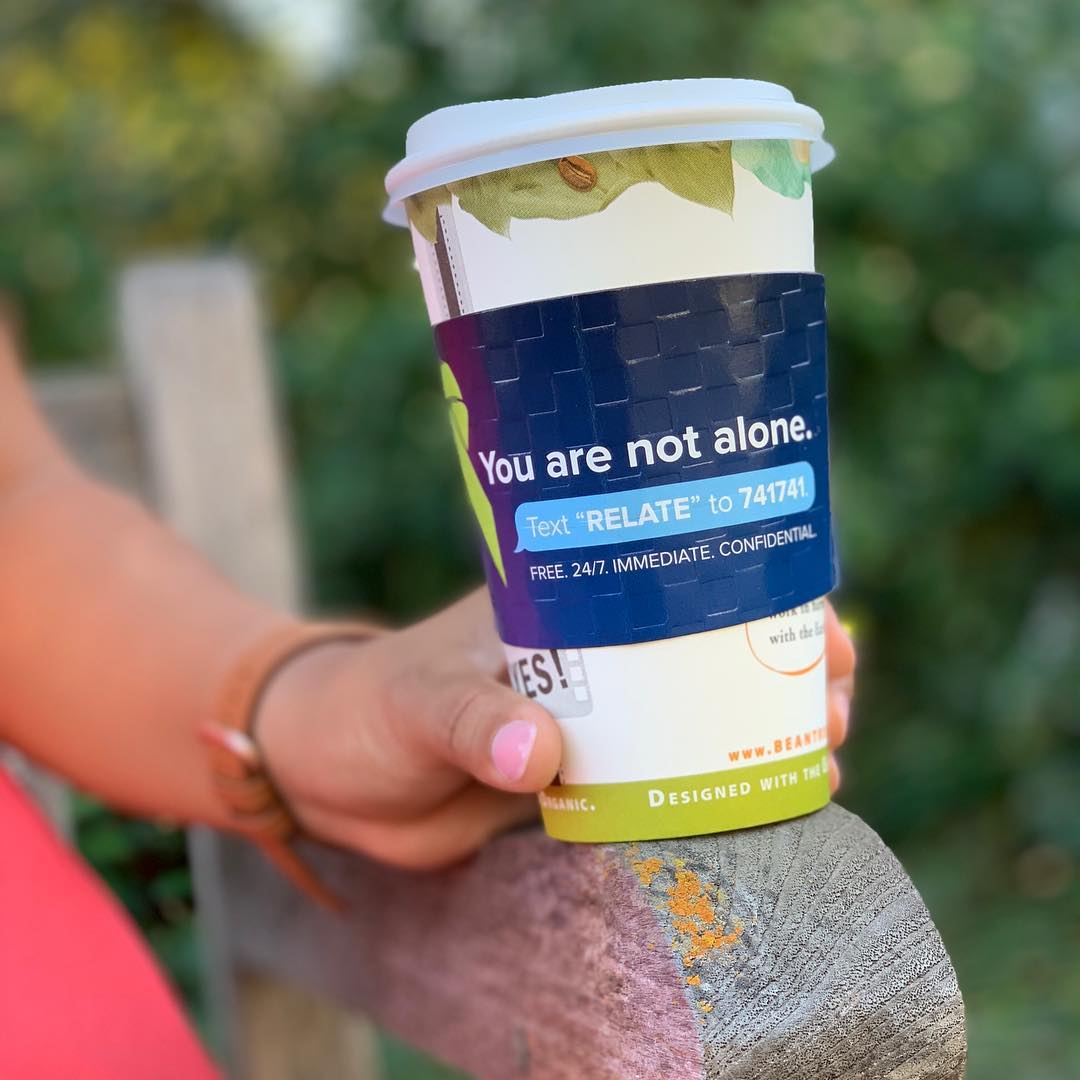 Person holding coffee cup with a Crisis Text Line coffee sleeve that reads: "You are not alone. Text "RELATE" to 741741" Free 24/7. IMMEDIATE. CONFIDENTIAL.