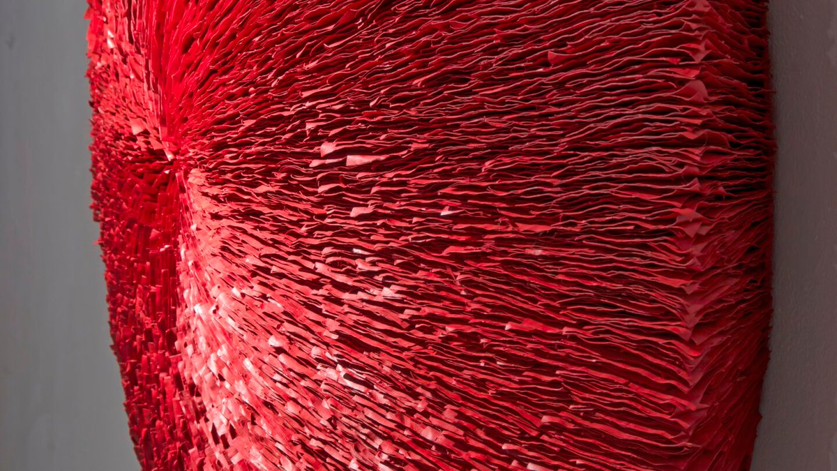 An oblique view of a deep red abstract round wall sculpture made from stacks of paper. Surface is highly textured with lines radiating outwards from the center.