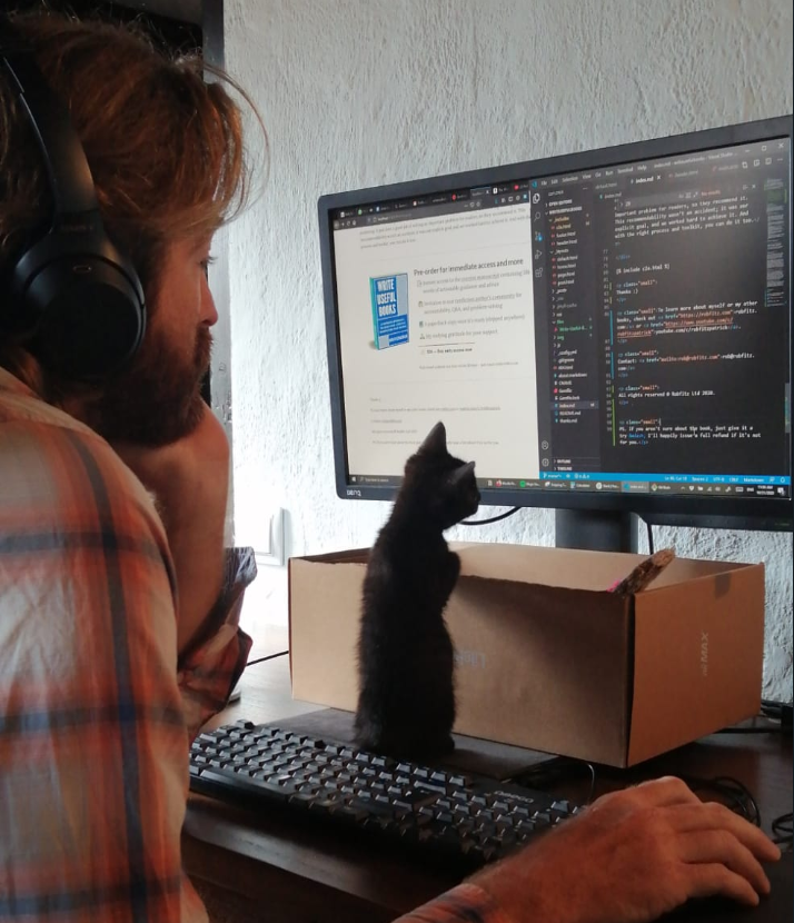 A kitten (named Couscous) standing on its hind legs between my keyboard and monitor, watching the screen as I code and debug the website for my new book.