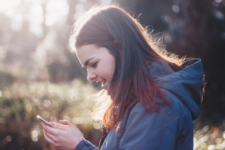 a young woman looking down at her smarphone smiling, wearing a jacket with a hoodie