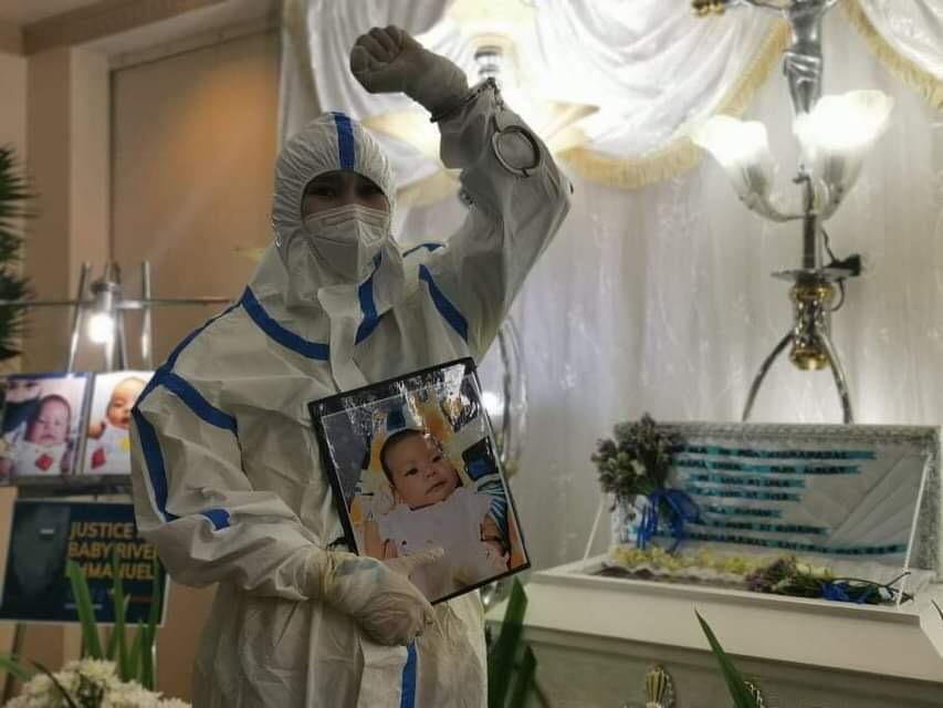 Handcuffs finally removed as Nasino holds a picture of baby River on her right hand and raises a clenched fist on the left. She is wearing a full PPE suit.  KAPATID