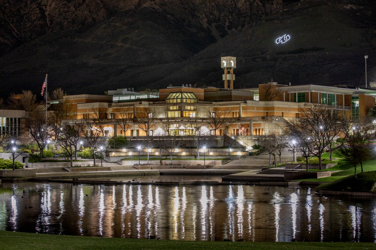 A photo of Weber State University's campus from 2020. The campus is illuminated at night and reflected into the duck pond. The "W" is lit up on the mountainside. 