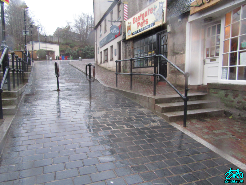 The barriers are normally locked across the route creating a chicane to slow down "whizzing" cyclists. But they can be unlocked & turned for parades etc and fully removed for emergencies. It should be noted that the barriers are across the road. It's footway that's lacking. 3/7