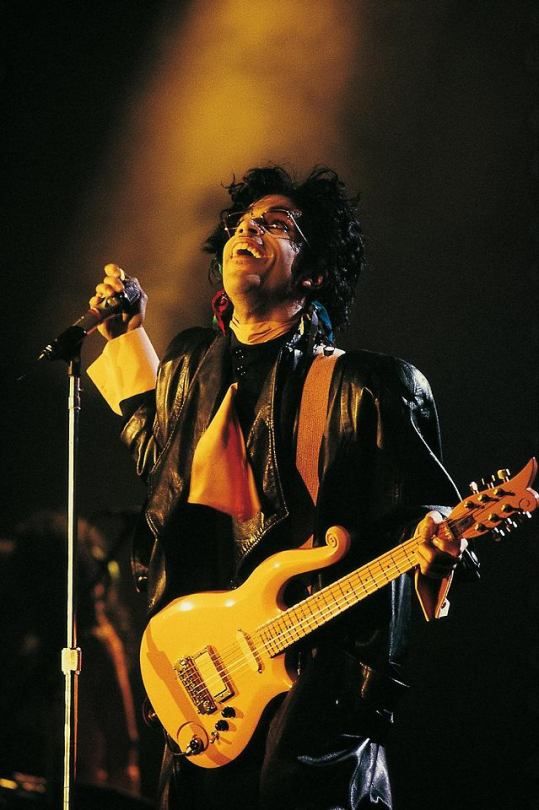 The guitar is the focus of the outfit, with the visible peach cuffs blending seamlessly with the instrument. As Prince performs, the coat flies open, flashing an all-peach silhouette, enrobed in high contrast black leather.
