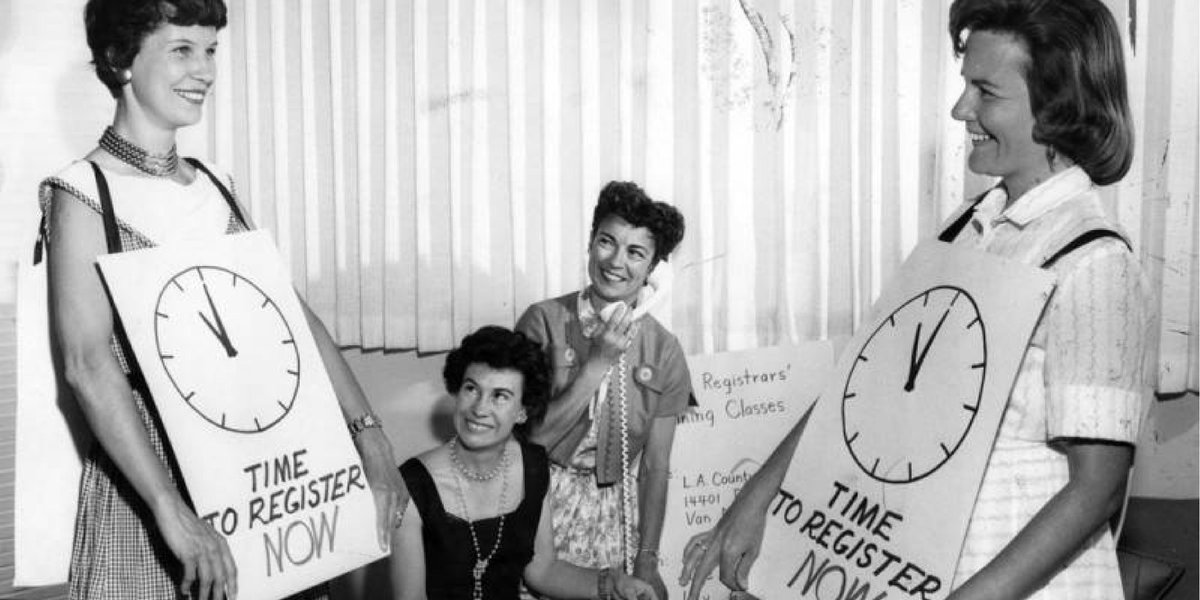 Black and white photo of two women holding signs of a clock that reads "time to register now" with two women in the background