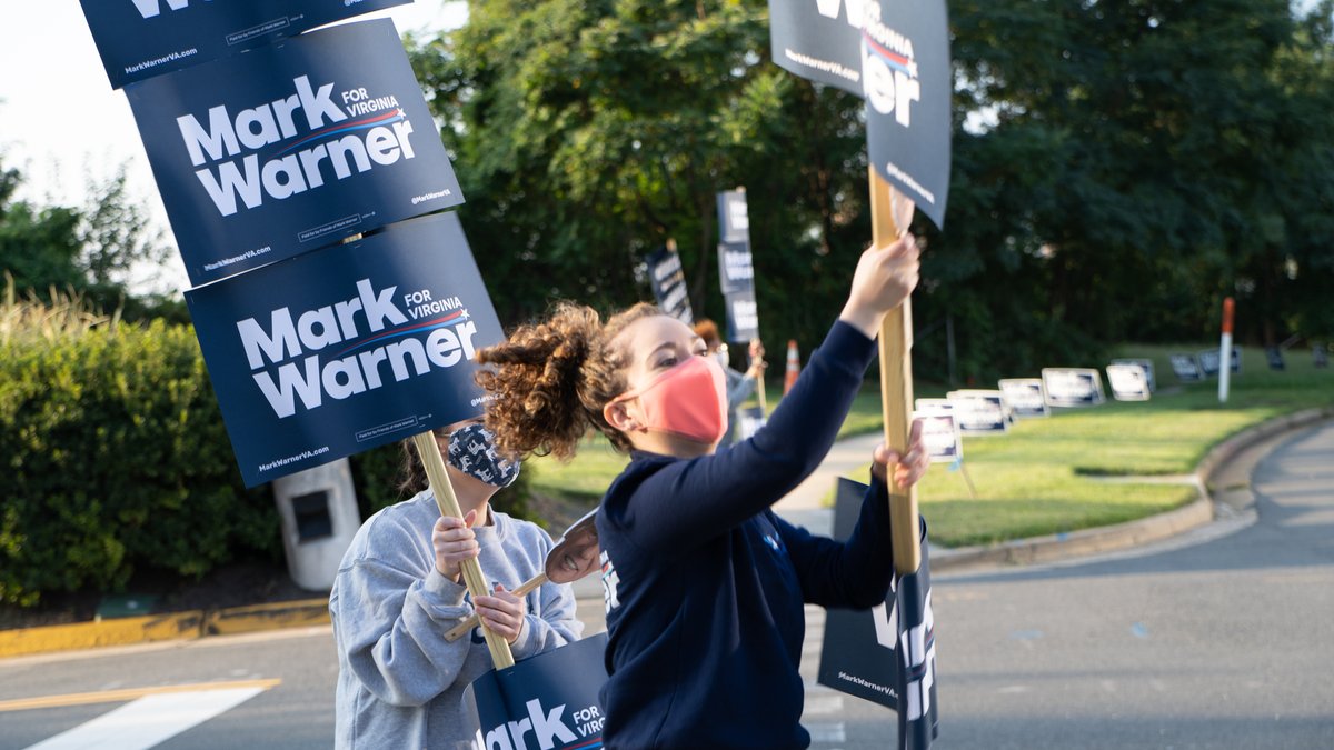 Two people stand outside holding signs that say "Mark Warner for Virginia."