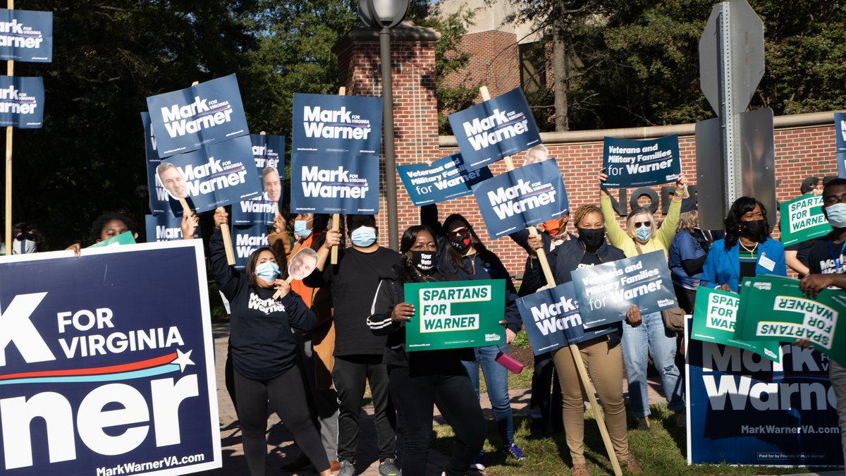 A crowd of people stand outdoors with signs that say "Mark Warner for Virginia" and "Spartans for Warner."