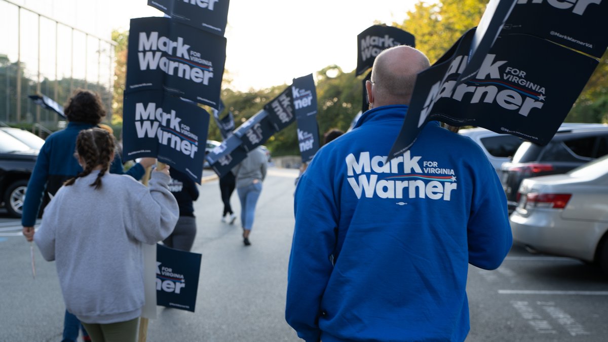 A crowd of people face away from the camera, holding signs that say "Mark Warner for Virginia."