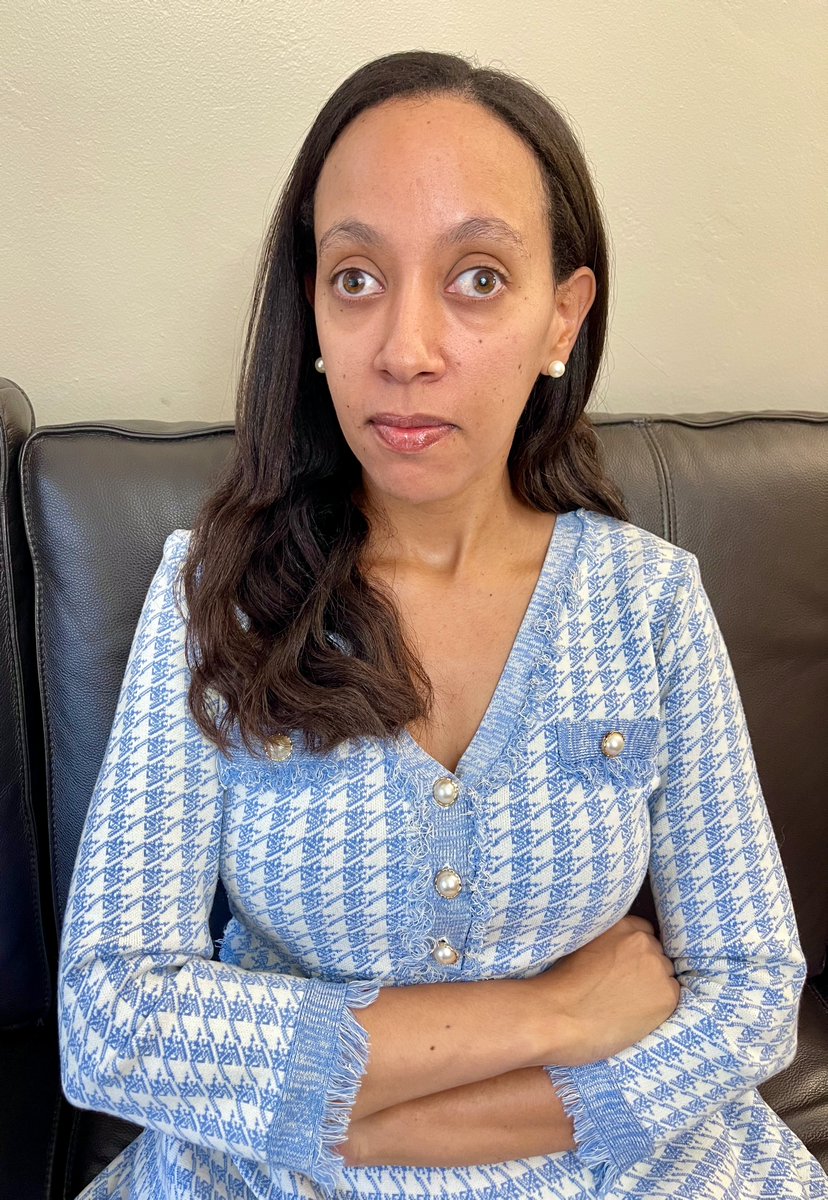 I'm sitting with my arms crossed on a black sofa. I'm wearing a blue dress and a serious expression.