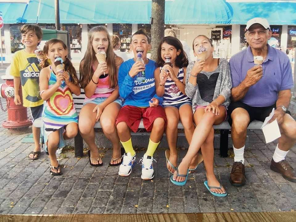 Representative Khan's 6 young grandchildren sit on a bench eating ice cream. At the end of the bench on the right side sits her late husband, Nasir Khan. 