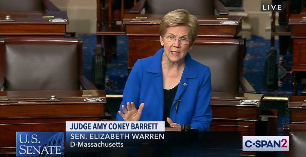 A screenshot of Elizabeth Warren, a white presenting woman with glasses, a blue jacket and a black shirt, in a Congressional room on CSPAN
