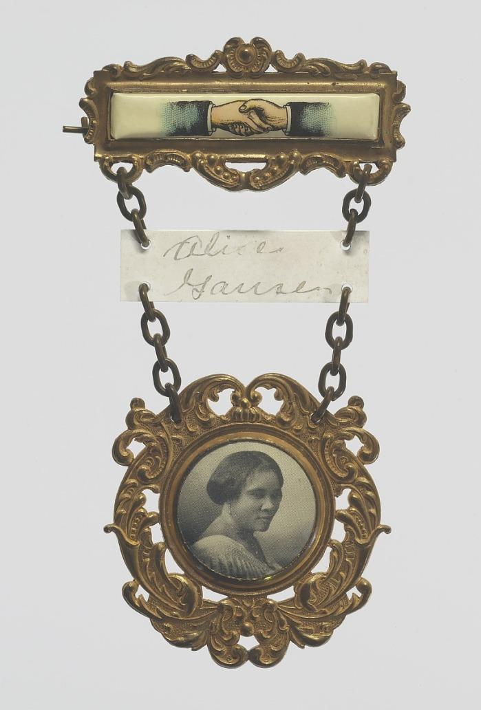 Badge from Madam C.J. Walker convention. It shows two hands shaking as though in a business deal. In a circular frame with decorative flourishes around it, a portrait of Madam C. J. Walker.
