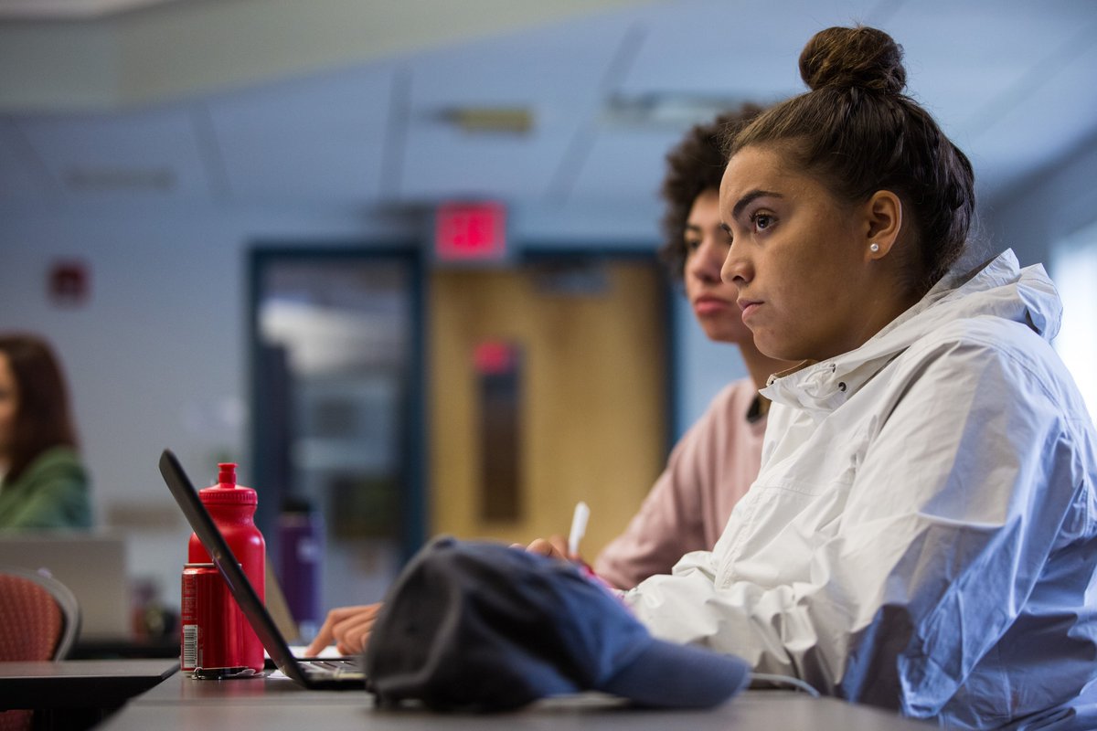 Two students sit in a classroom. One typing on a laptop.