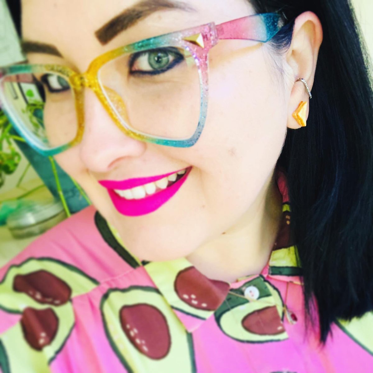 Selfie of me (pale skin, dark hair) smiling. I’m wearing cheese toastie shaped earrings and a pink dress with avocado print.