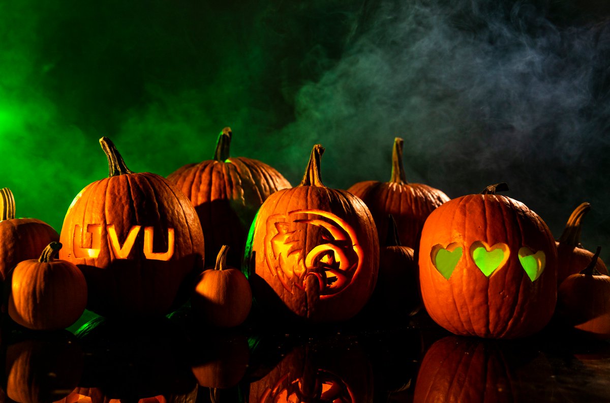 Three carved pumpkins — UVU monogram, wolverine head, and three green hearts — surrounded by other pumpkins of various sizes. Green and white fog swirling in the background to give a mysterious effect. 