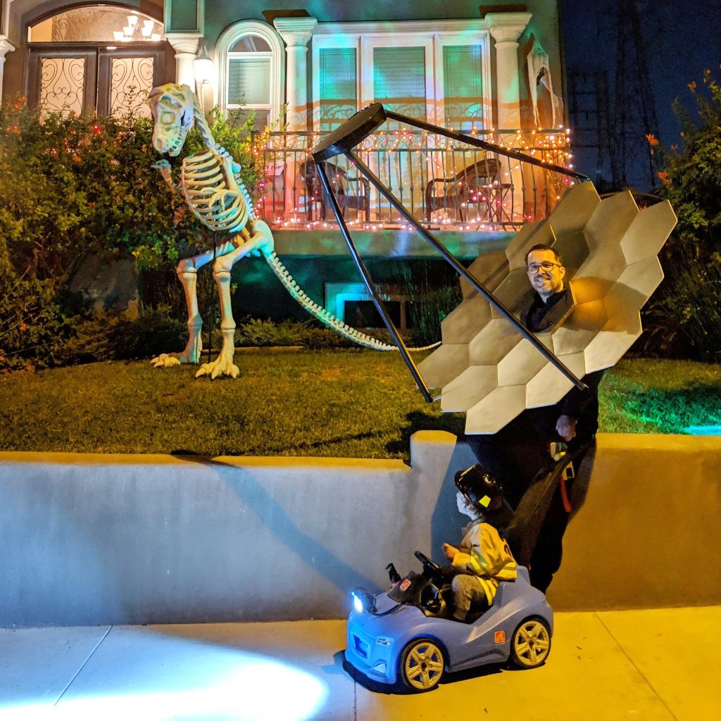Tony Long in a James Webb Space Telescope primary mirror costume, with his child in a small toy car besides him. In the background there is a giant dinosaur skeleton.