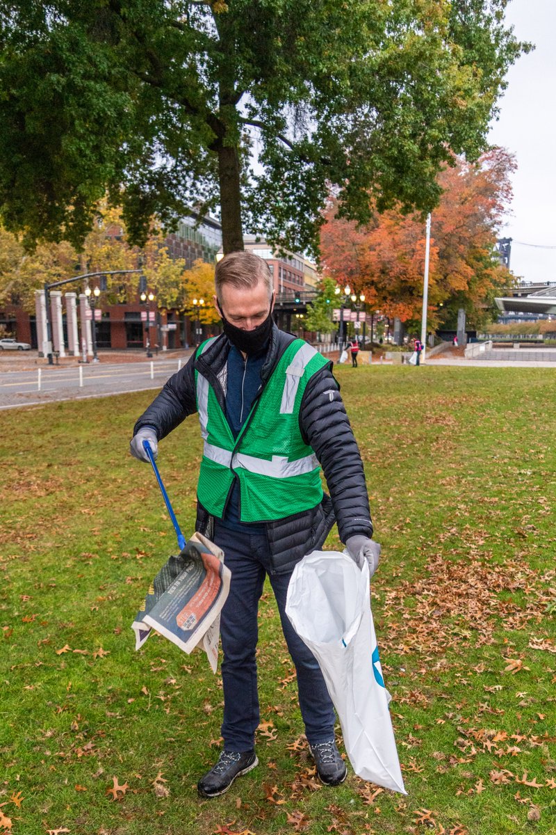 Mayor Ted Wheeler stands in Waterfront Park in Portland. He is wearing a face mask and a reflective vest and is putting a piece of litter into a trash bag using a trash picker.