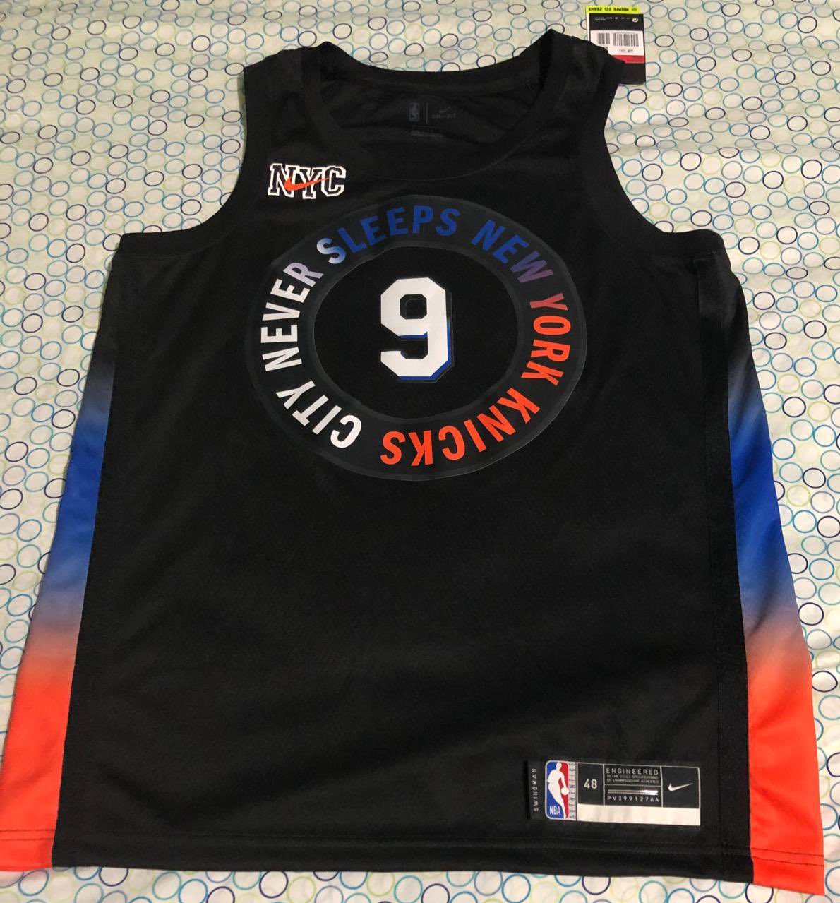 Chemise noire des New York Knicks, avec des détails bleus et orange sur les côtés et le logo sur la poitrine qui dit "CITY NEVER SLEEPS NEW YORK KNICKS".  Le logo Nike a les lettres NYC au dos.  La chemise numéro 9 est sur un lit et le couvre-lit a des motifs à pois.