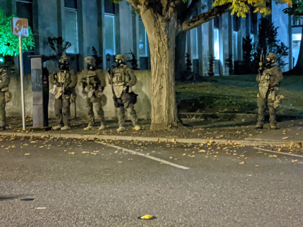 Four people in military uniforms, including ridiculous crotch armor, stand in front of a short wall. Behind the wall, a building with long rectangular windows