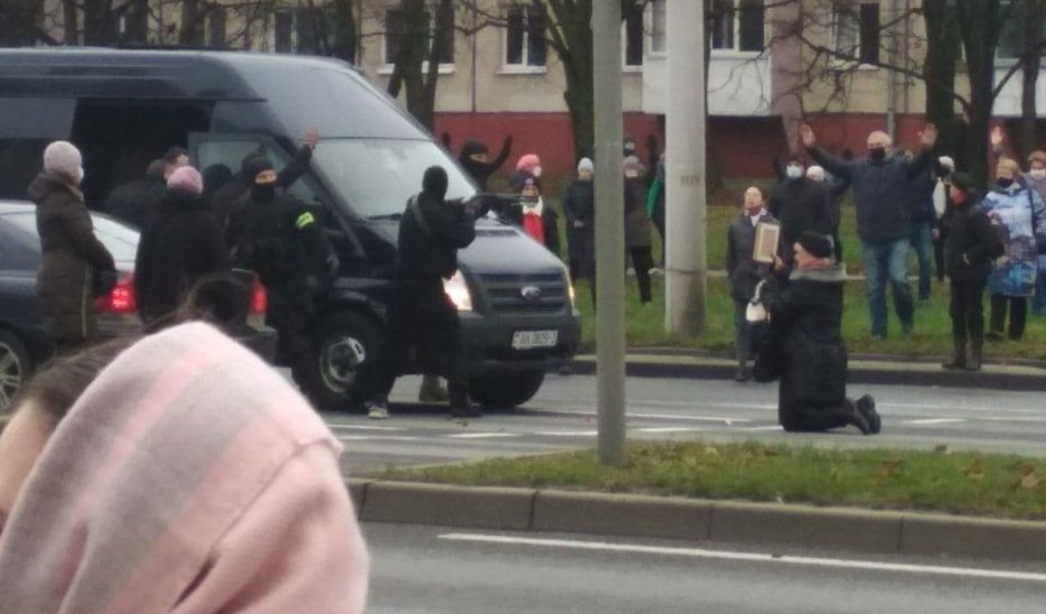 A woman is kneeling in front of a van while a masked man points a gun at her