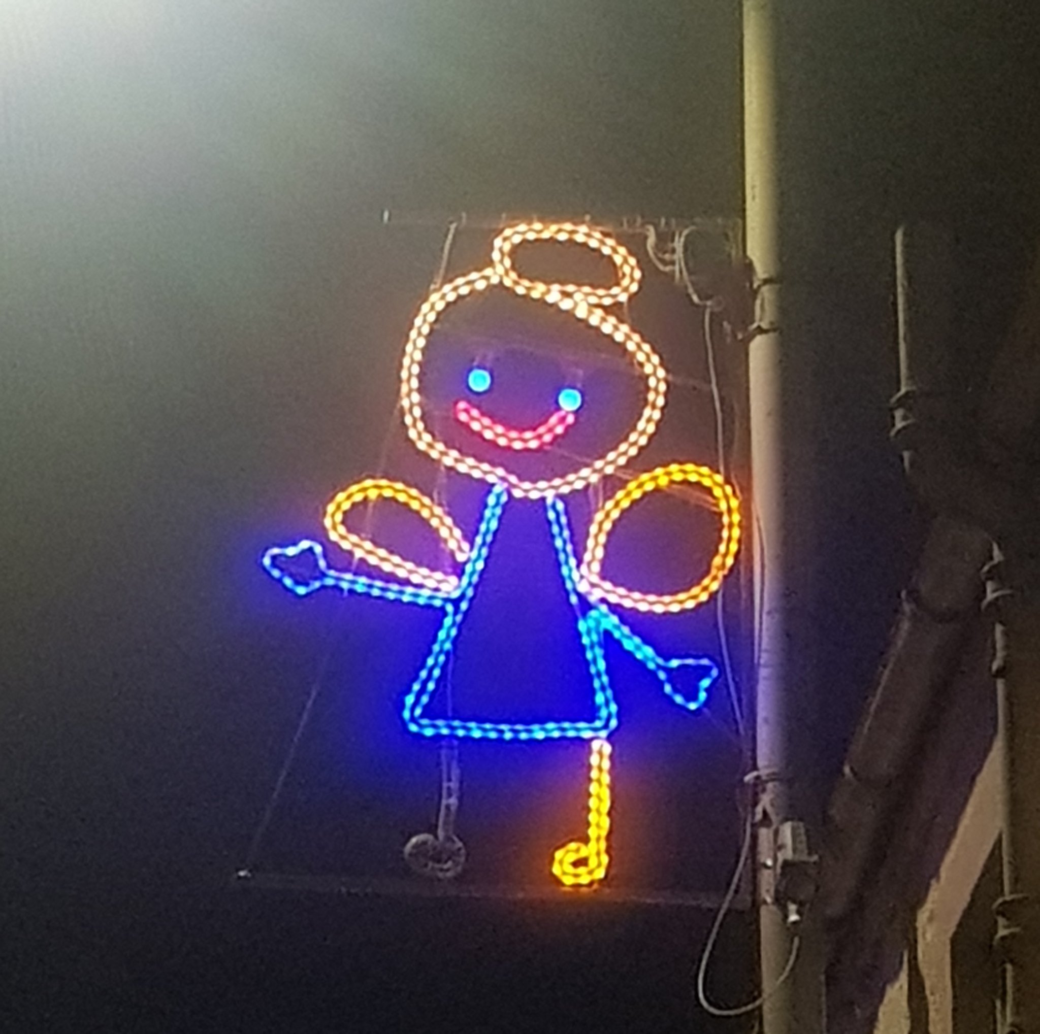 A photo of a city's Christmas lights on a light pole, in the shape of a child's drawing of an angel
