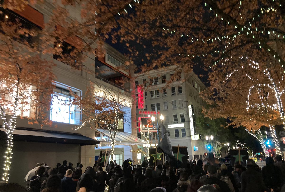 Hundreds march through downtown Portland under trees turning orange for Fall covered in twinkling lights.
