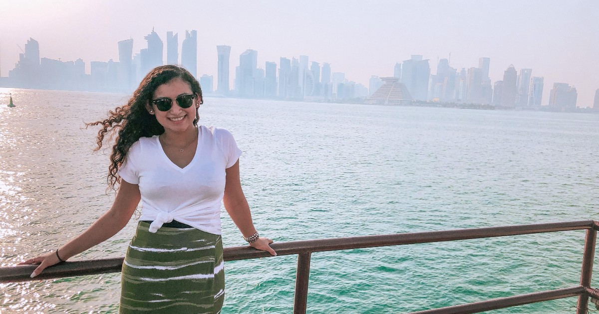 Outdoors, image of a female student standing by a railing in front of a city skyline and body of water, wearing white shirt, green skirt, and sunglasses