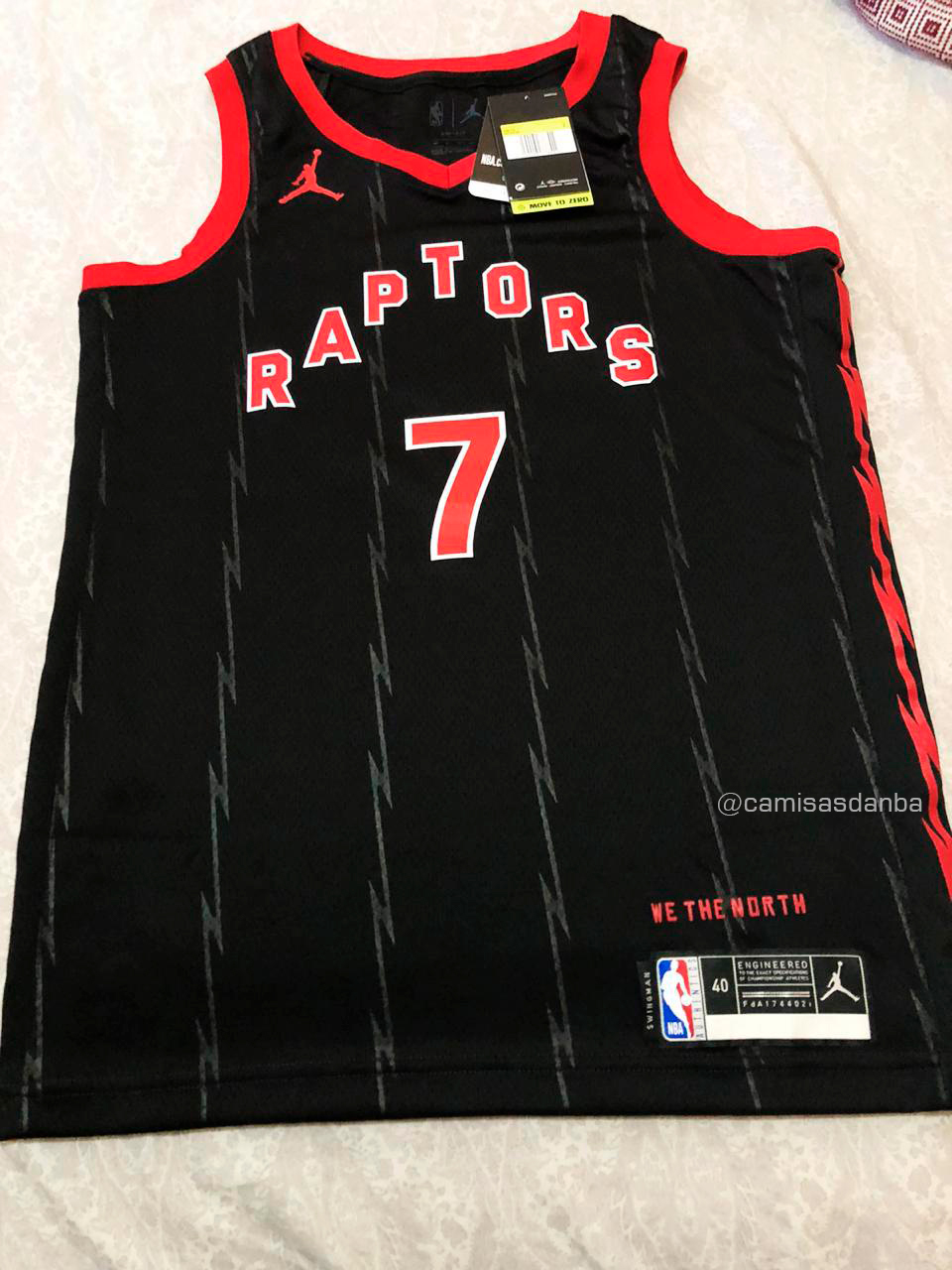 Camisa preta do Toronto Raptors sobre uma cama. A camisa tem seis linhas verticais finas em cinza escuro brilhante que forma um ziguezague. Nas laterais as linhas aparecem um pouco mais grossas, mas na cor vermelho. Ela tem gola e mangas em vermelho, o logo da Jordan Brand vermelho próximo ao ombro direito, as letras da palavra RAPTORS em formato de arco com ângulo agudo no peito, apontando para cima. Elas são vermelhas com contorno branco, assim como o número 7 abaixo delas. Próximo à etiqueta da barra da camisa está a frase "We The North" (algo como "nós somos o norte", em tradução livre). A imagem tem uma marca d'água do perfil Camisas da NBA.