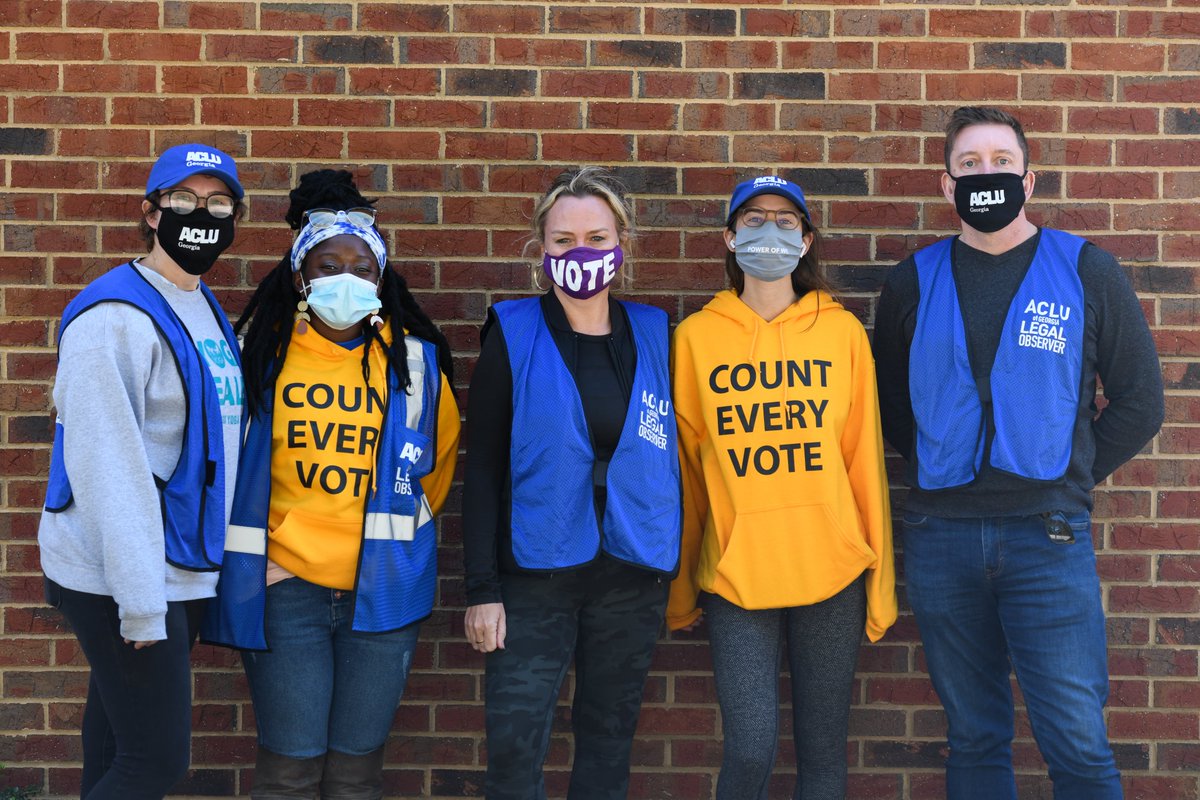Horizontal photo of multiple poll workers standing in front of a brick wall. All are wearing blue vests that say "ACLU LEGAL OBSERVER" and masks, some that read VOTE and others "ACLU of Georgia." 2 of the poll workers are wearing sweatshirts that say "COUNT EVERY VOTE." They are all looking toward the camera.