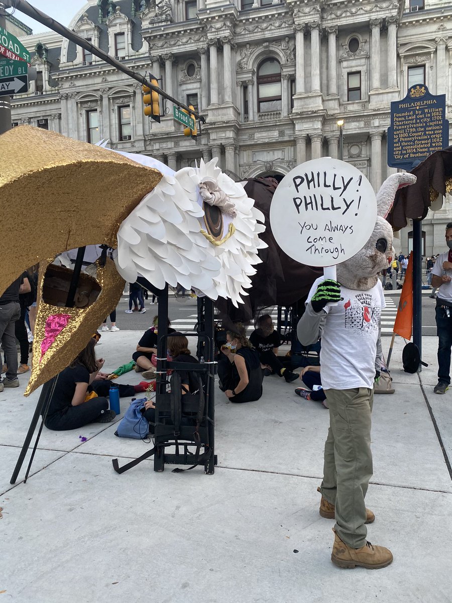 a rabbit in front of a big eagle in front of Philly city hall holding a sign that reads “philly! philly! you always come through!”